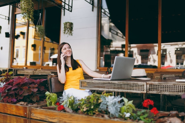 Happy girl in outdoors street cafe sitting at table with laptop pc computer, talking on mobile phone, conducting pleasant conversation, in restaurant during free time