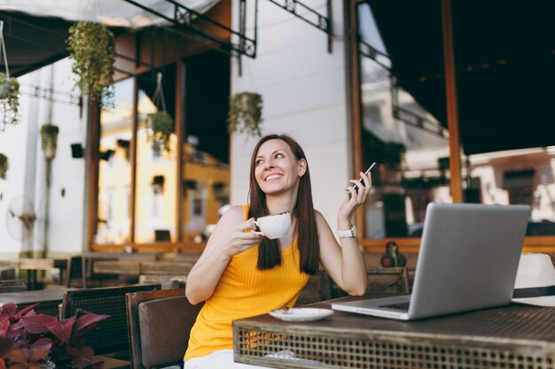 Happy girl in outdoors street coffee shop cafe sitting at table with laptop pc computer, texting message on mobile phone, drink cup tea in restaurant during free time