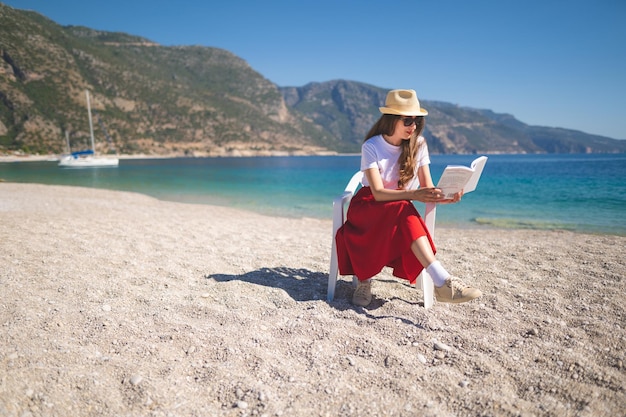 Happy girl in a red skirt on an empty beach sits on a chair and reads a book