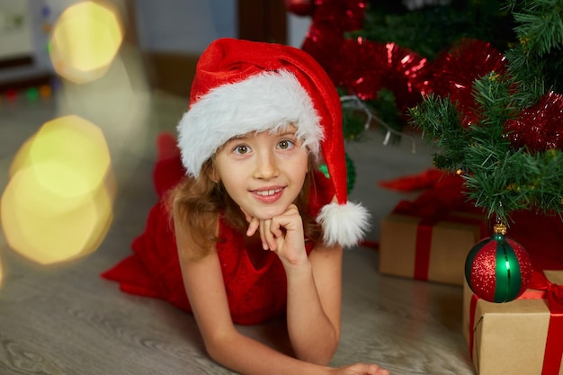 Happy Girl in Santa hat lying near christmas tree at home