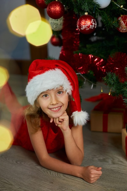 Happy Girl in Santa hat lying near christmas tree at home