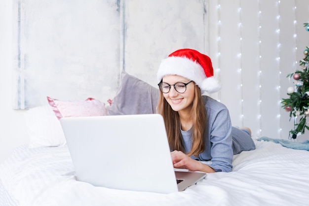 Happy girl in santa hat shopping online on laptop and sitting on bed next