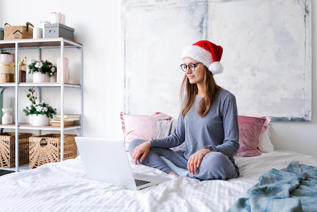 Happy girl in santa hat shopping online on laptop and sitting on bed next