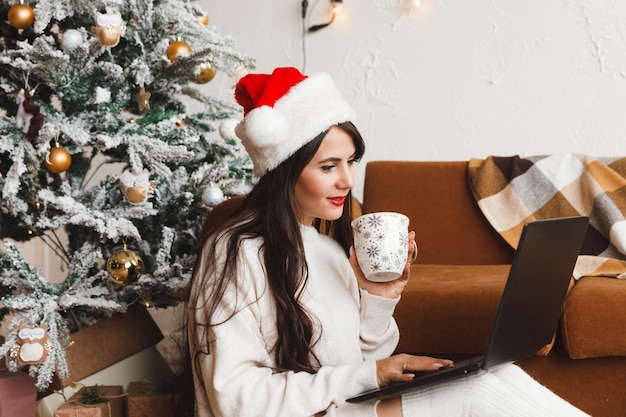 A happy girl in a Santa hat wishes her friends Merry Christmas in a video chat on a laptop A young woman lying under a Christmas tree among gift boxes in a home interior