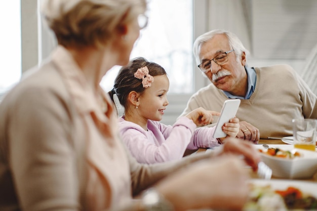 Happy girl showing something on mobile phone to her grandfather during a meal at dining table