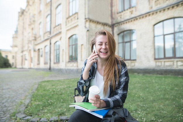 Happy girl sitting on a bench with books and coffee, talking on the phone and laughing.