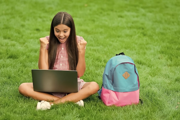 Happy girl sitting on green grass with laptop Start up child playing computer game back to school education online knowledge day kid learning private lesson blogging