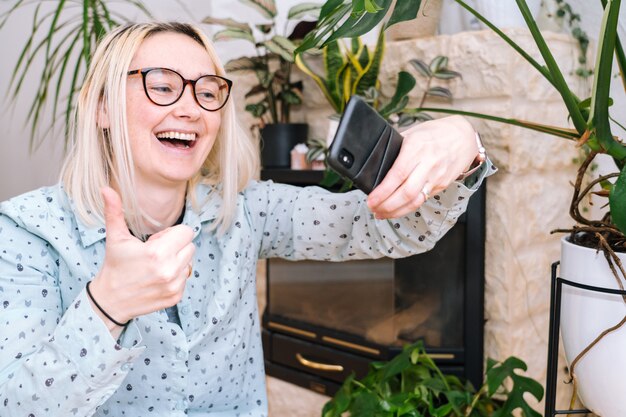 Photo happy girl sitting at home kitchen and holding videocall. young woman using smartphone for video call with friend or family. vlogger recording webinar. woman looking camera and waving greeting hands