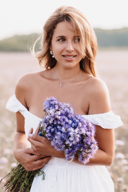 Happy girl smiles in a white dress with a bouquet of flowers in the field