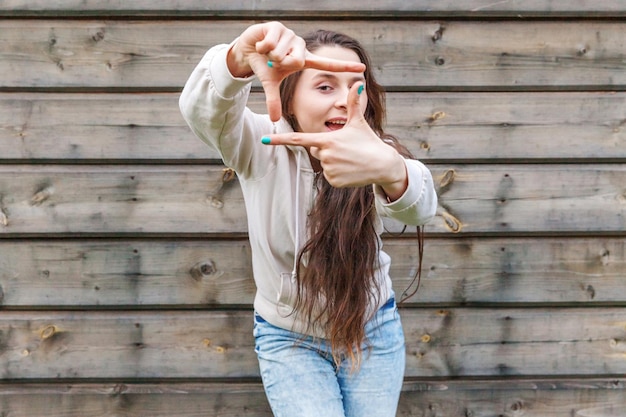 Happy girl smiling. Beauty portrait young laughing brunette woman showing frame with fingers on wooden wall background. European woman. Positive human emotion facial expression body language