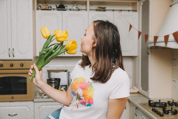 Happy girl stands in the kitchen with a bouquet of yellow tulips flowers for birthday mothers day easter