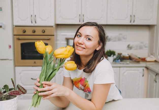 Happy girl stands in the kitchen with a bouquet of yellow tulips flowers for birthday mothers day easter