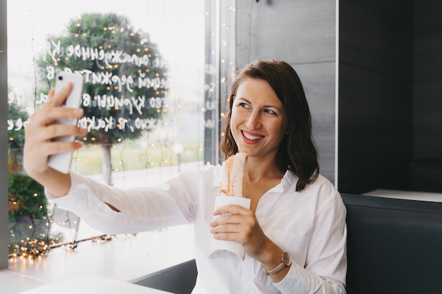 Happy girl takes a selfie on a mobile phone while eating a twister in a cafe.