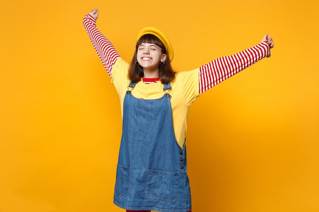 Happy girl teenager in french beret, denim sundress keeping eyes closed, spreading and rising hands isolated on yellow wall background. People sincere emotions, lifestyle concept. Mock up copy space.