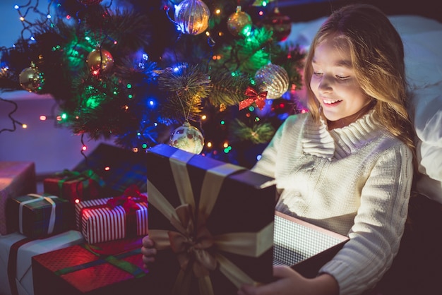 The happy girl unpack a gift box on a christmas tree background. night time