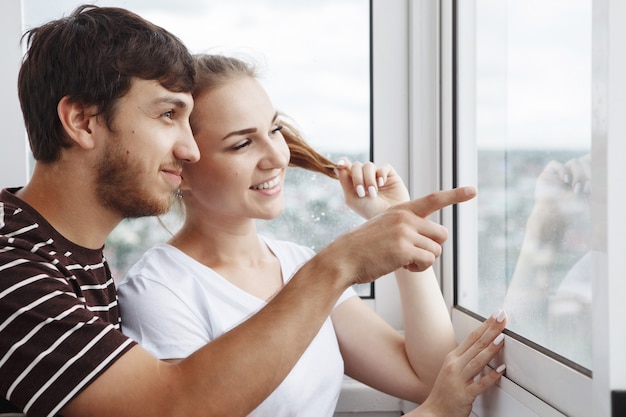 Happy girl in white t-shirt and guy with a beard, cute couple looking out the window in their new home