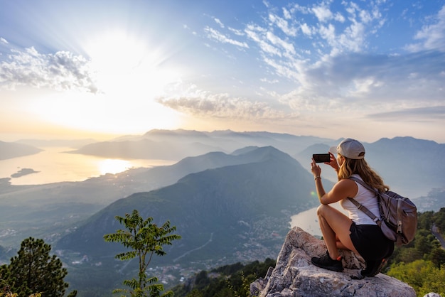 A happy girl with a backpack photographs the seascapes of montenegro from the top of the mountain