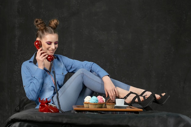 Happy girl with bright makeup and bun hairstyle holds retro phone Black background maffins with multicolored cream