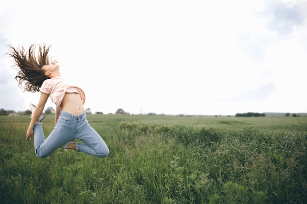 Happy girl with curly hair jumps on the green field against cloudy sky