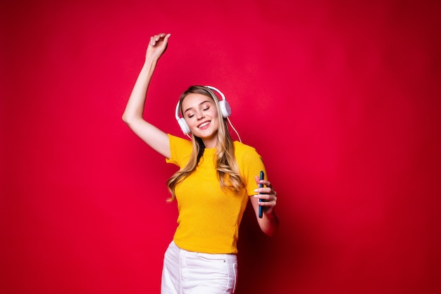 Happy girl with, listening to music in headphones and dancing, holding a smartphone. On a red wall.