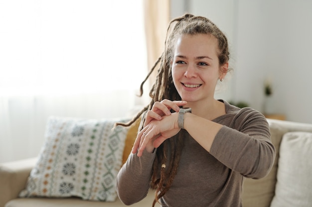 Happy girl with toothy smile keeping hand with smartwatch by her face while relaxing on couch at home