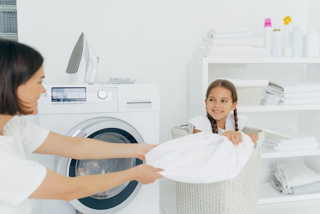 Happy girl with two pigtails poses in basket with dirty linen has fun in laundry room with mother helps to do washing Woman loads washing machine spend weekend at home busy with domestic work