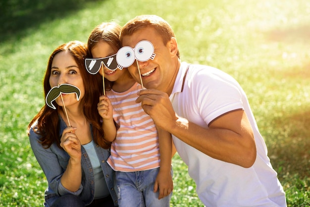 Photo happy girl woman and man using funny paper masks on the sticks in the park and looking at the camera