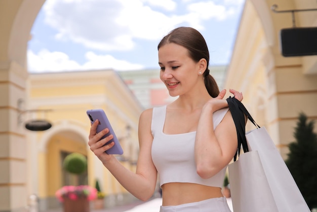 Happy girl young woman is smile looking at screen of smartphone In mall carrying shopping paper bag