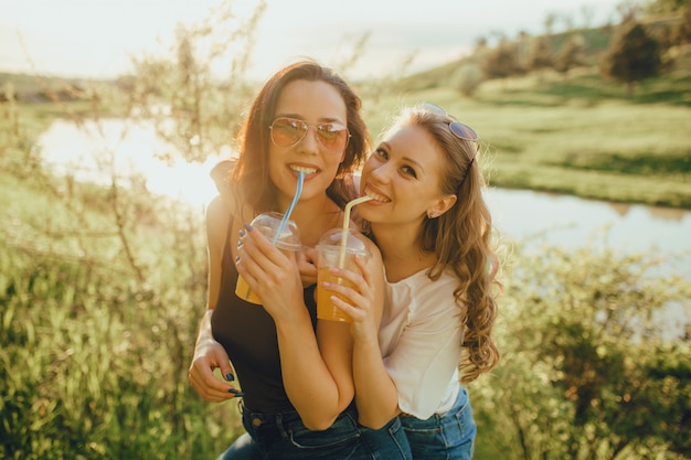 Happy girls have fun, drink cocktail from plastic cup with straw in sunglasses, dressed in white and black shirt, at sunset, positive facial expression, outdoor