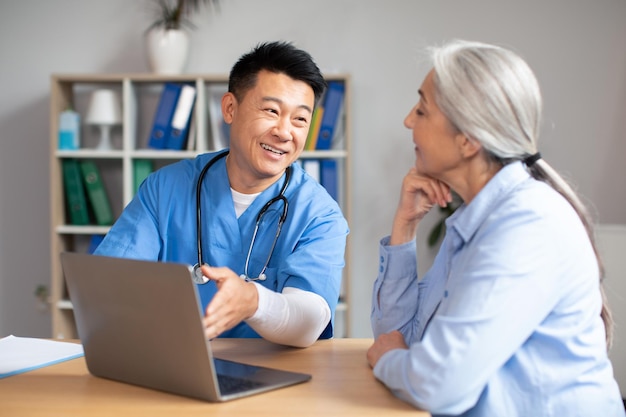 Happy glad handsome middle aged korean doctor shows laptop to old female caucasian patient in clinic