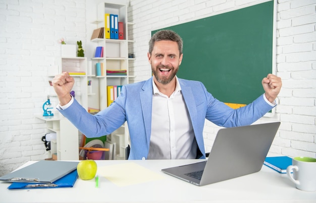 Happy glad school teacher in classroom with computer at blackboard