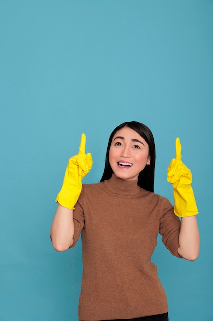 Happy glade and cheerful asian woman pointing fingers upward to copy space Isolated on a blue background, Housewife worker, Cleaning home concept, Smiling joyful female with positive state of mind