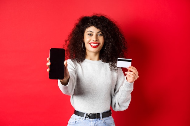 Happy good-looking girl showing plastic credit card and empty smartphone screen, demonstrate account or application, standing on red background