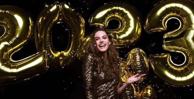 Happy gorgeous girl in stylish sexy party dress holding gold 2023 balloons having fun at New Year's Eve Party