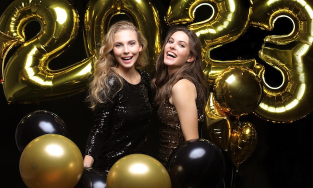 Happy gorgeous girls in stylish sexy party dresses holding gold 2023 balloons having fun at New Year's Eve Party