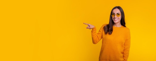 Happy gorgeous young brunette woman in a sweater and sunglasses is pointing away while she posing isolated on yellow background