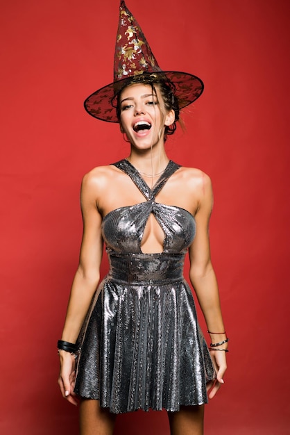 Happy gothic young woman in witch halloween costume with hat standing and smiling over red background Pretty young blonde woman dressed as a fairy Attractive model girl in Halloween costume