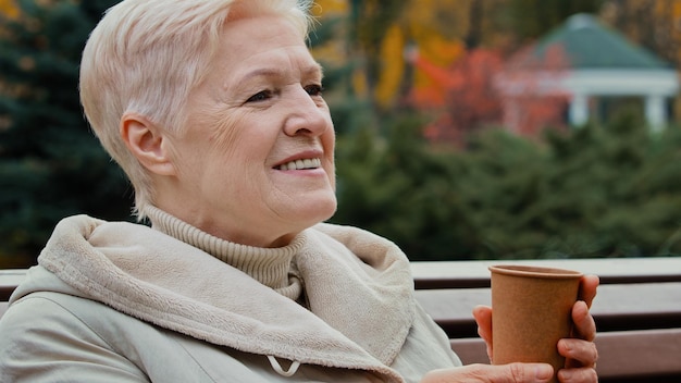 Happy grayhaired dreamful mature elderly woman grandmother drinking coffee hot tea from disposable