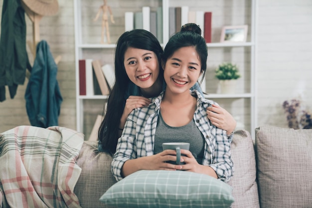 Happy group of beautiful young asian female friends sitting arm in arm on sofa in living room smiling face at camera at home. girls sisters hugging lovely. woman weekend vacation holiday concept