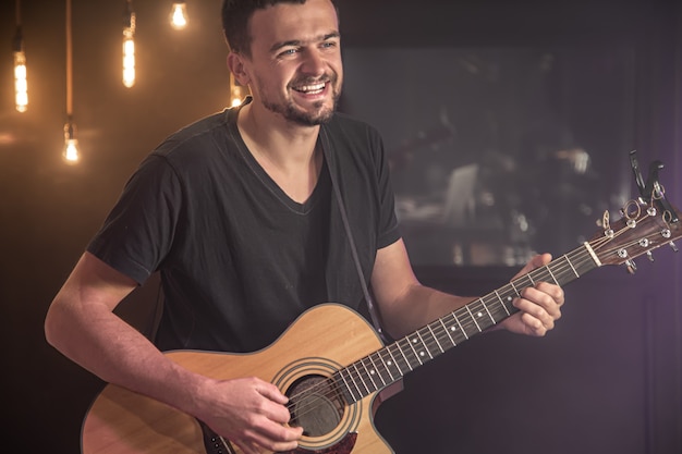 Happy guitarist in a black t-shirt plays an acoustic guitar at a concert against a blurred black background.