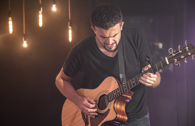 Happy guitarist in a black t-shirt plays an acoustic guitar at a concert against a blurred black wall.