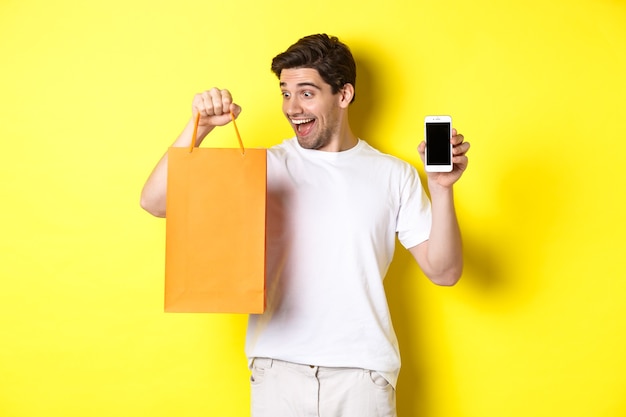 Happy guy buy something in store and looking at shopping bag, showing mobile phone screen, yellow wall