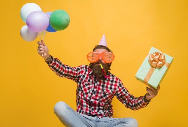 Happy guy celebrating birthday cheerful bearded man in holiday cap with balloons and present box