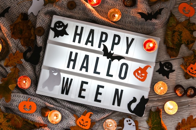 HAPPY HALLOWEEN. Text in white light box, with a background of sweater, dried leaves halloween autumn decoration. Ghost, pumpkins bat, black cat. Home decoration. Top view. Bokeh light