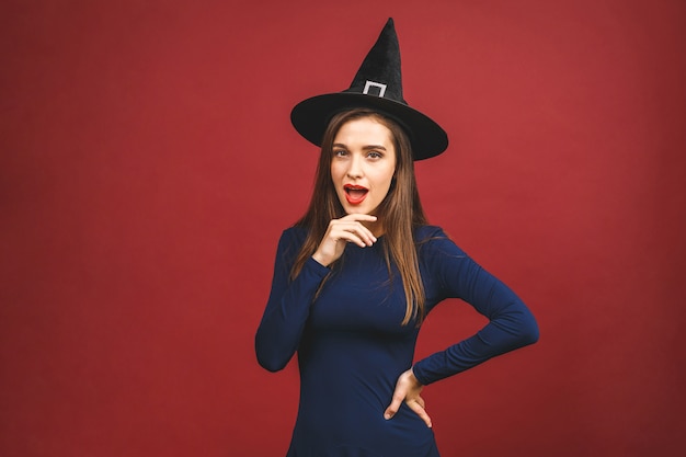 Happy Halloween Witch with bright make-up and long hair. Beautiful young surprised woman posing in witches sexy costume. Isolated on red background.