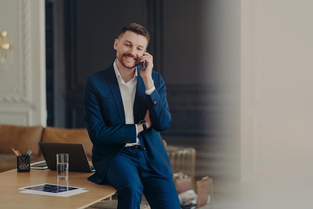 Happy handsome businessman talking on phone while sitting on office desk