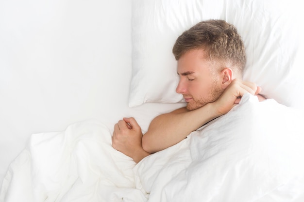 Happy handsome guy, young man is sleeping, having good dreams, smiling, lying on white pillow, covering with blanket. Copy space, top view. Healthy well sleep, daily regime, relaxation, rest concept.