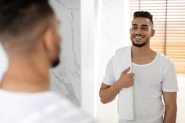 Happy handsome middle eastern man holding towel and smiling to mirror