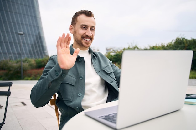 Happy handsome modern man waving hand to laptop web camera at street cafe