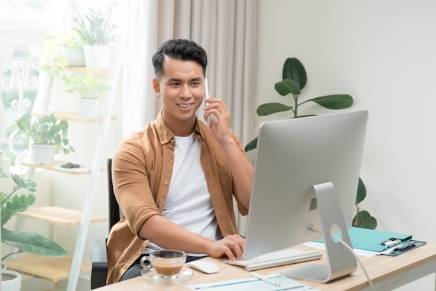 Happy handsome young businessman using computer and talking on cell phone in office